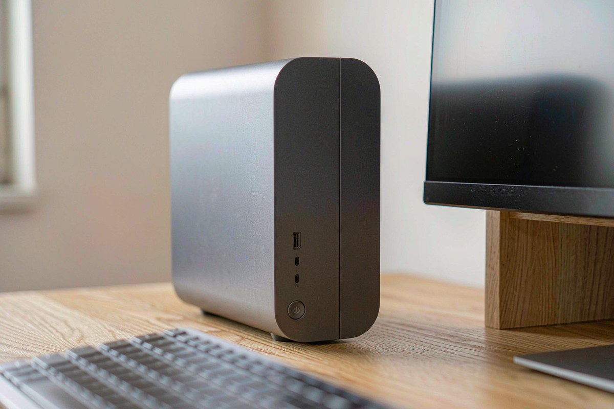 A compact silver ACEMAGIC micro desktop PC standing on a light oak desk beside a monitor