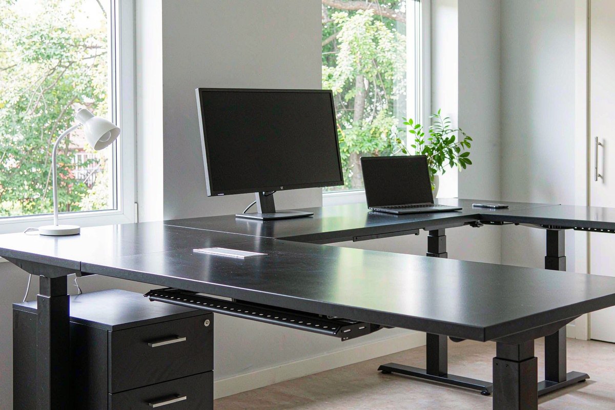 A large black L-shaped height-adjustable standing desk with drawers in a bright corner home office