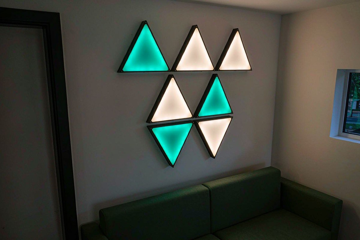 Seven triangular Nanoleaf Shapes panels glowing teal and warm white on a white wall behind a green sofa